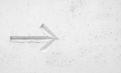 A white, textured concrete wall with a carved arrow pointing left, showing subtle shadows and cracks.